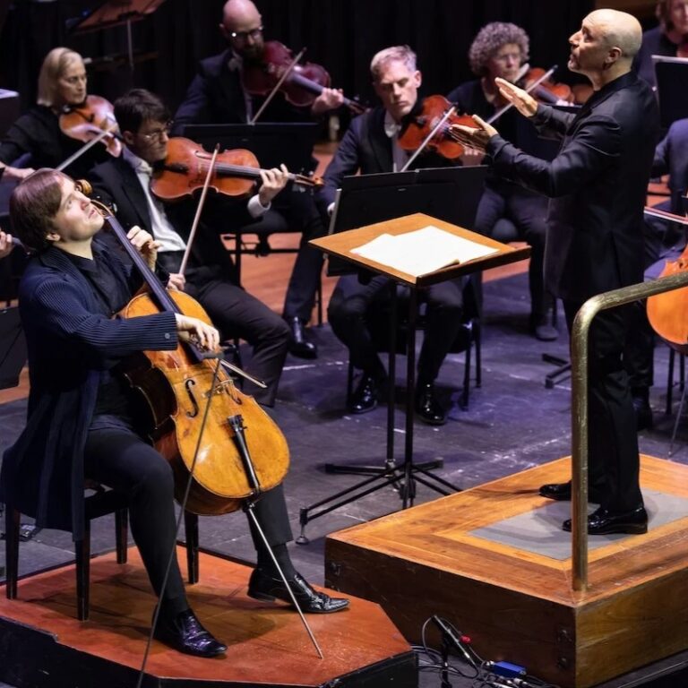 A man playing a cello in front of a symphony orchestra as the conductor directs nearby.